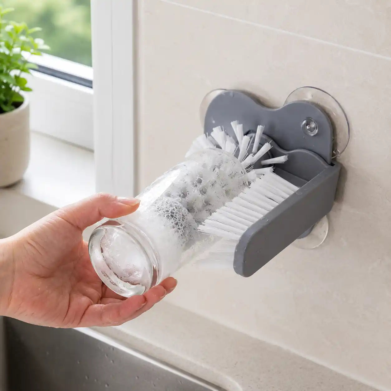 Lifestyle image of a grey 2-in-1 suction cup glass cleaning brush attached beside a modern kitchen sink, showing a drinking glass being cleaned with dual-sided white bristles for fast and efficient 360° cleaning.
