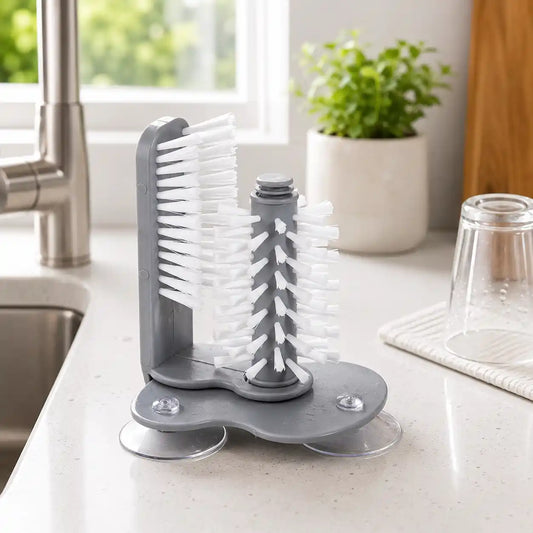 Lifestyle image of a grey 2-in-1 suction cup glass and bottle cleaning brush placed beside a modern kitchen sink, featuring white nylon bristles and a compact design for cleaning glasses, mugs, bottles, and drinkware.